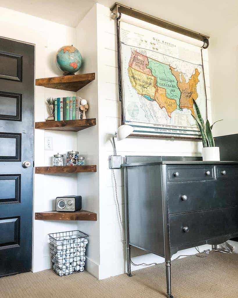 Rustic wooden corner floating shelves with books, a globe, and storage jars complement a vintage map and black dresser in a cozy space