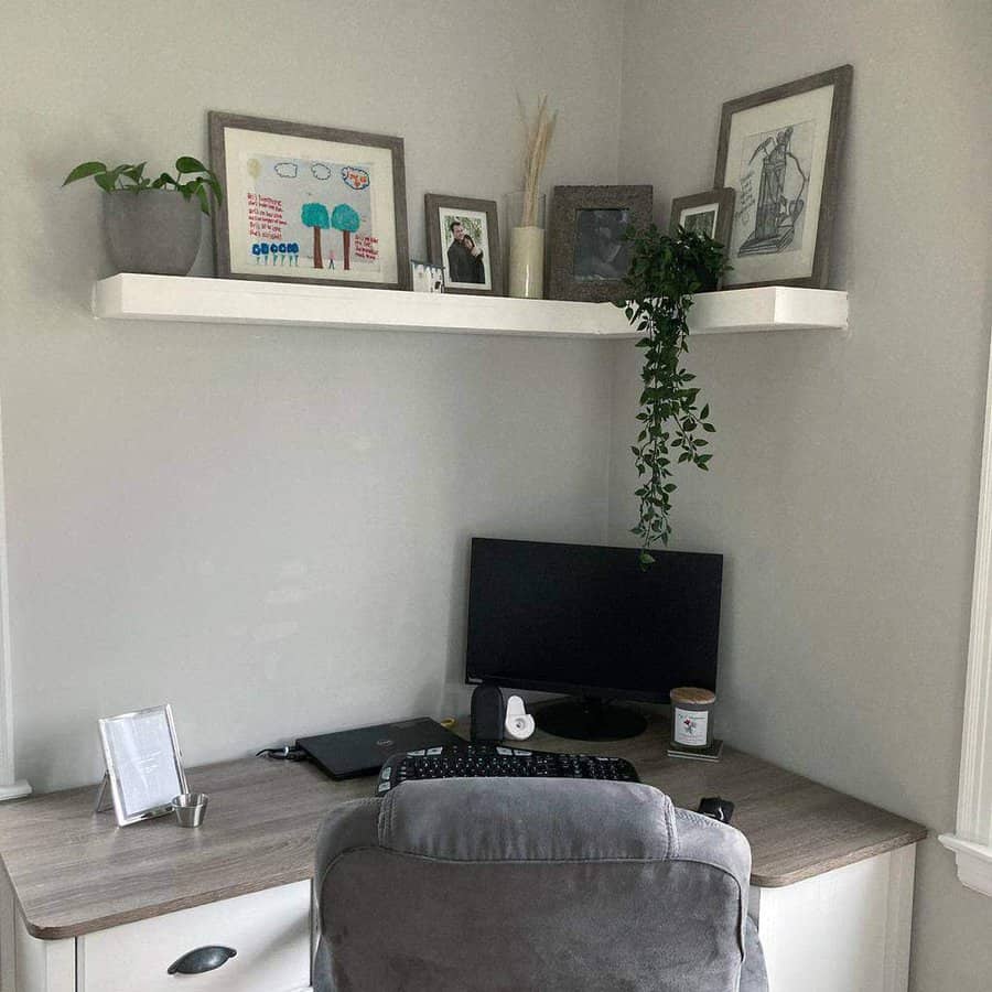 A cozy home office with a desk, computer, and chair; a white corner shelf above holds photos, plants, and art; walls are light gray