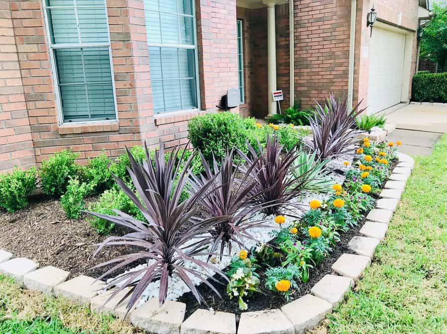 Front yard landscaping with colorful flowers, purple foliage plants, trimmed shrubs, and a stone border along a brick house