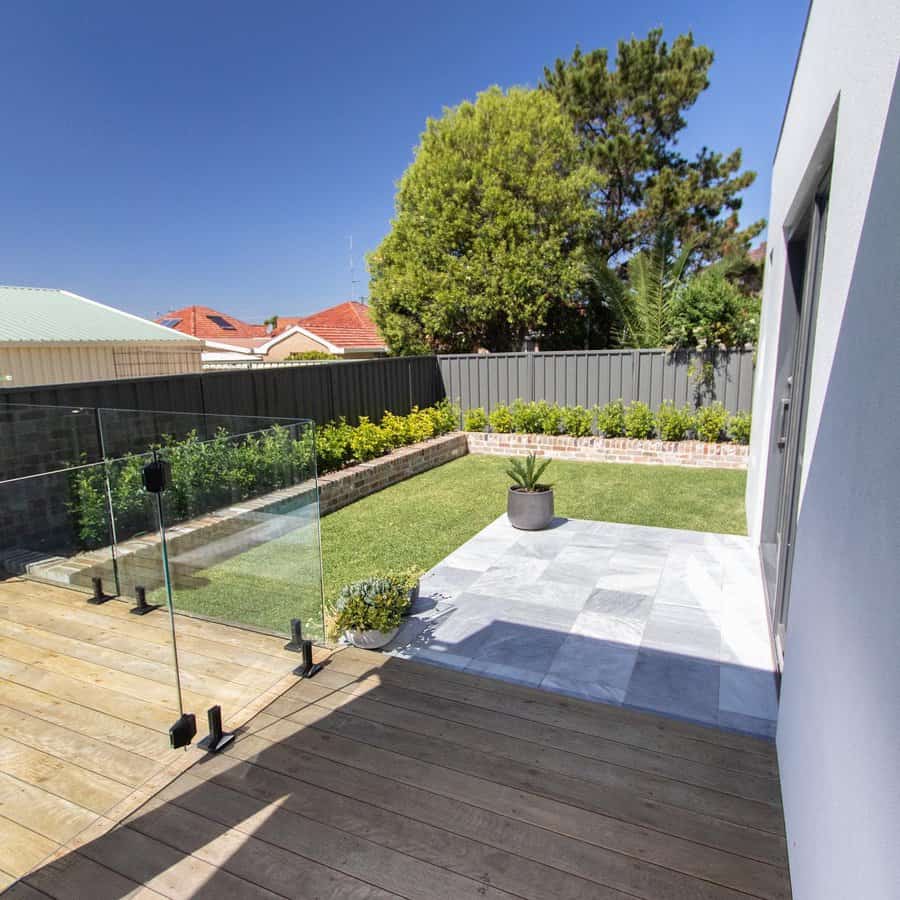 Modern backyard with a wooden deck, glass fence, neatly trimmed lawn, tiled patio area, and minimal landscaping for a clean look