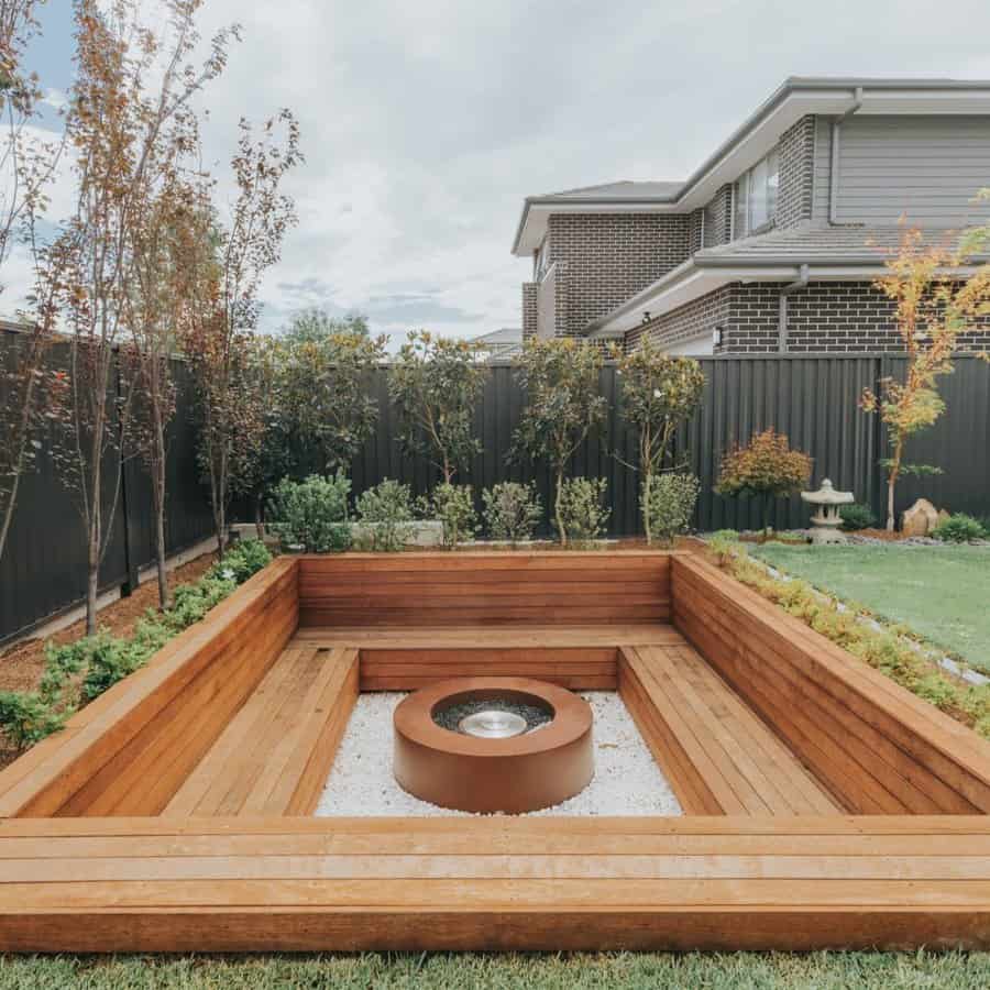 Cozy backyard fire pit area with a wooden seating bench, gravel base, lush greenery, and modern privacy fencing