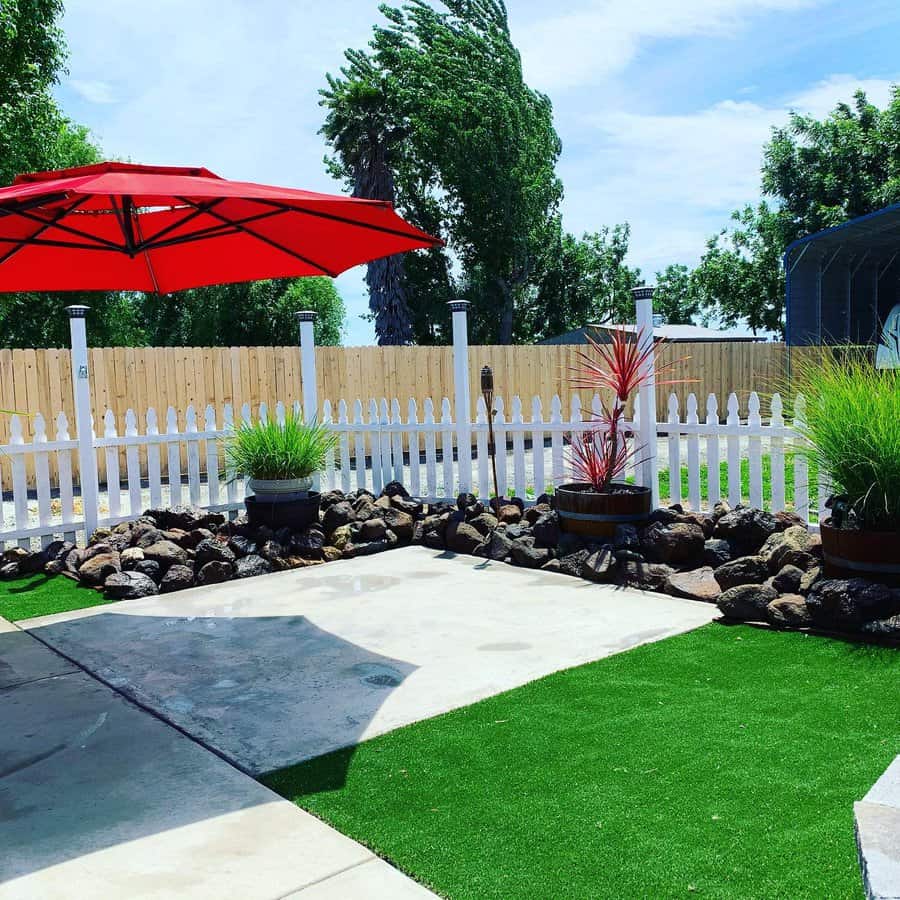 Bright backyard with artificial turf, a white picket fence, rock landscaping, potted plants, and a red umbrella for shade