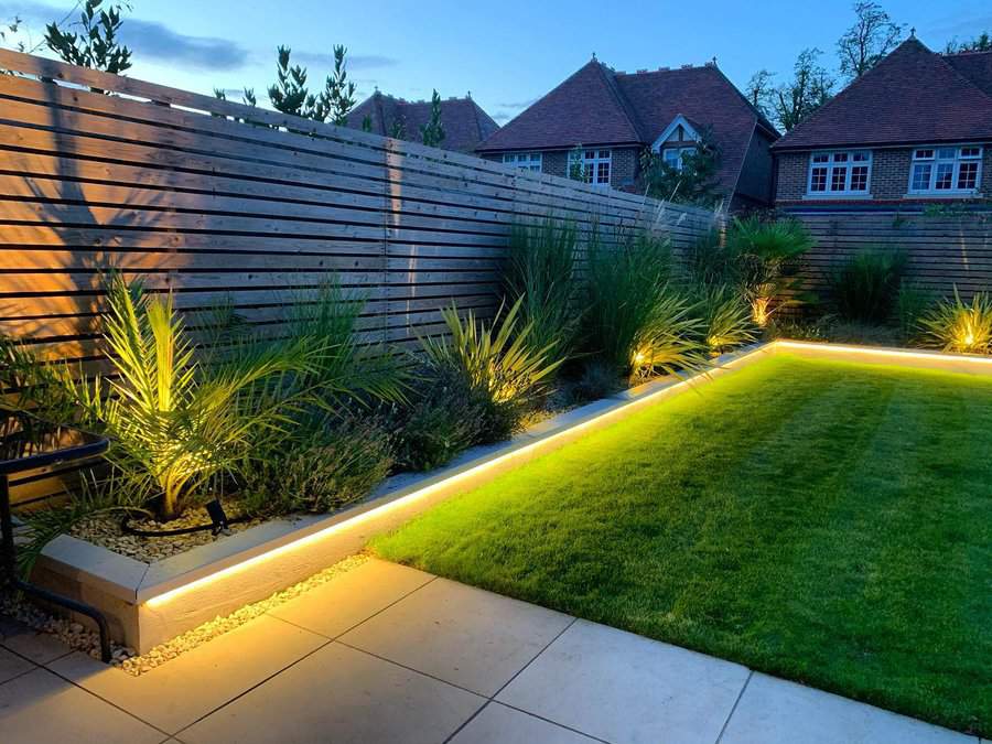 Modern backyard with wooden privacy fence, uplighting on plants, lush lawn, and sleek perimeter LED lighting for evening ambiance