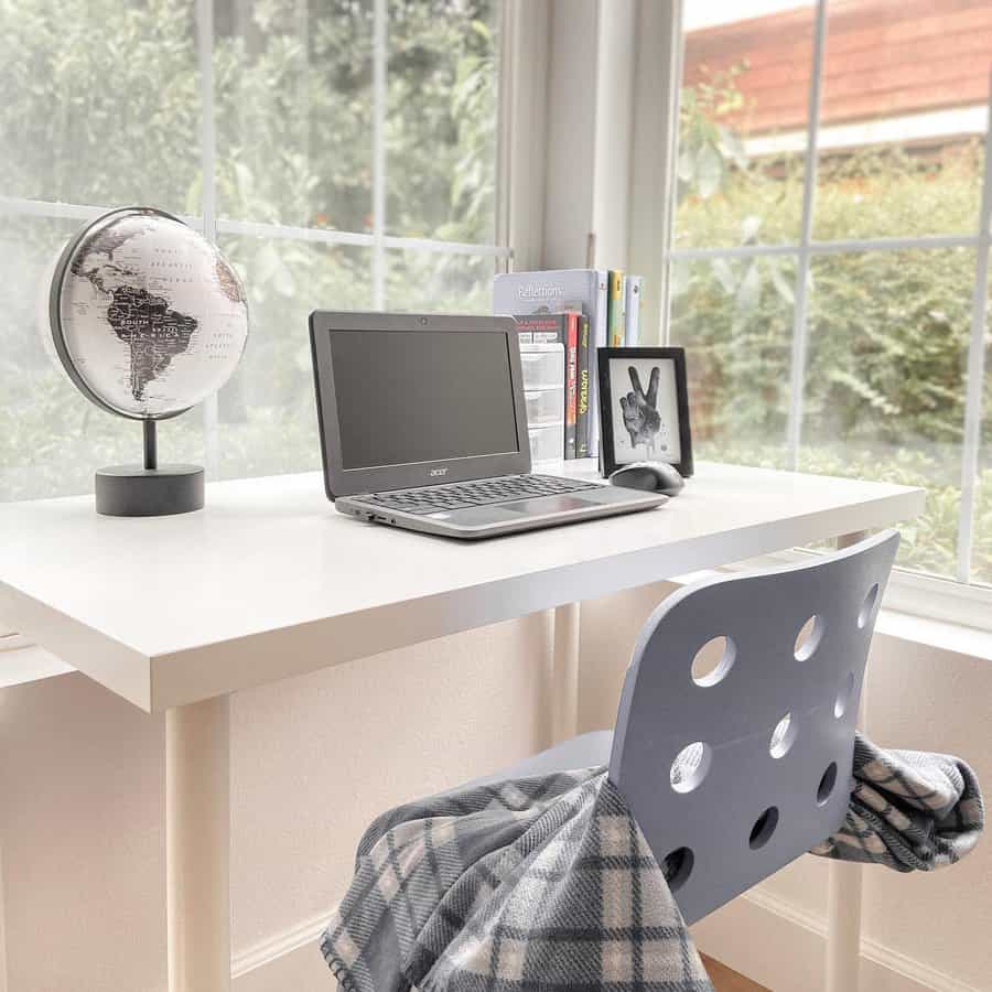 A minimalist workspace with a laptop, globe, books, and a framed picture on a white desk with a checkered blanket draped over a chair