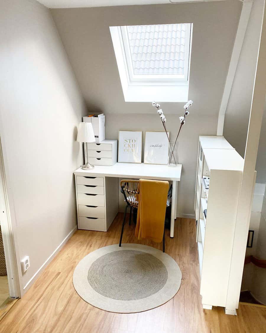 A small, cozy workspace with a white desk, chair, and drawers under a skylight featuring decor including a lamp, wall art, and a plant vase