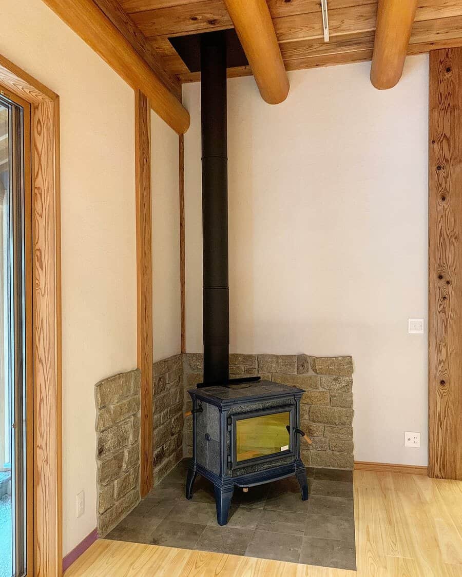 Rustic wood stove set in a stone-accented corner hearth, framed by exposed wooden beams and a tiled base for a cozy cabin feel.