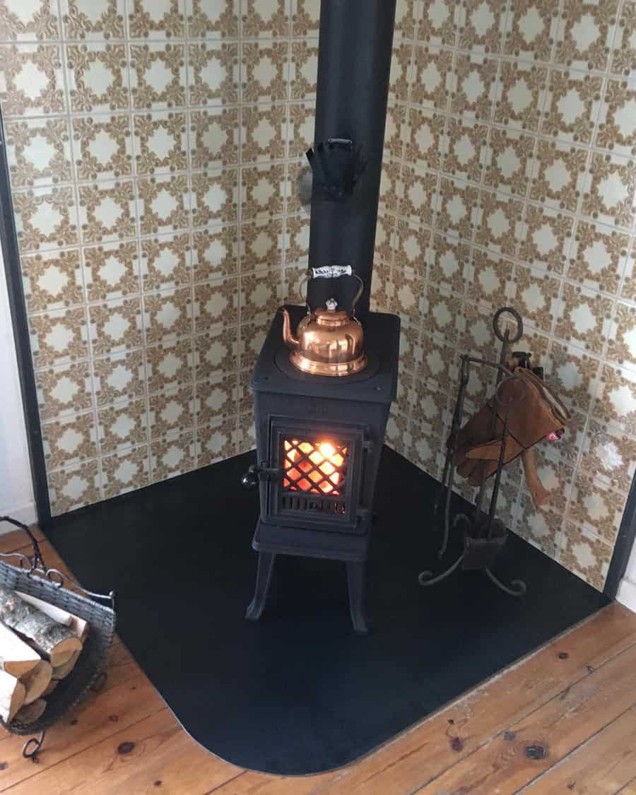 Compact wood stove in a vintage-style corner hearth with patterned tile walls, a black hearth pad, and a copper kettle for a cozy touch.