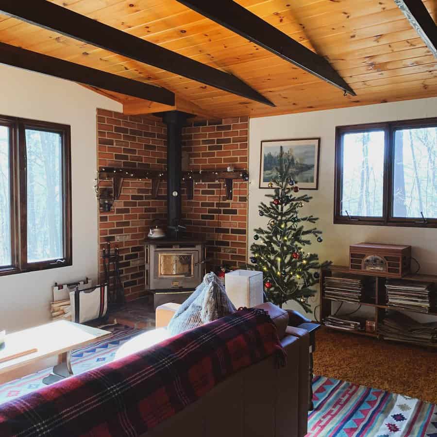 Cozy cabin-style living room with a brick corner wood stove, warm wooden ceiling beams, and a decorated Christmas tree for a festive touch.