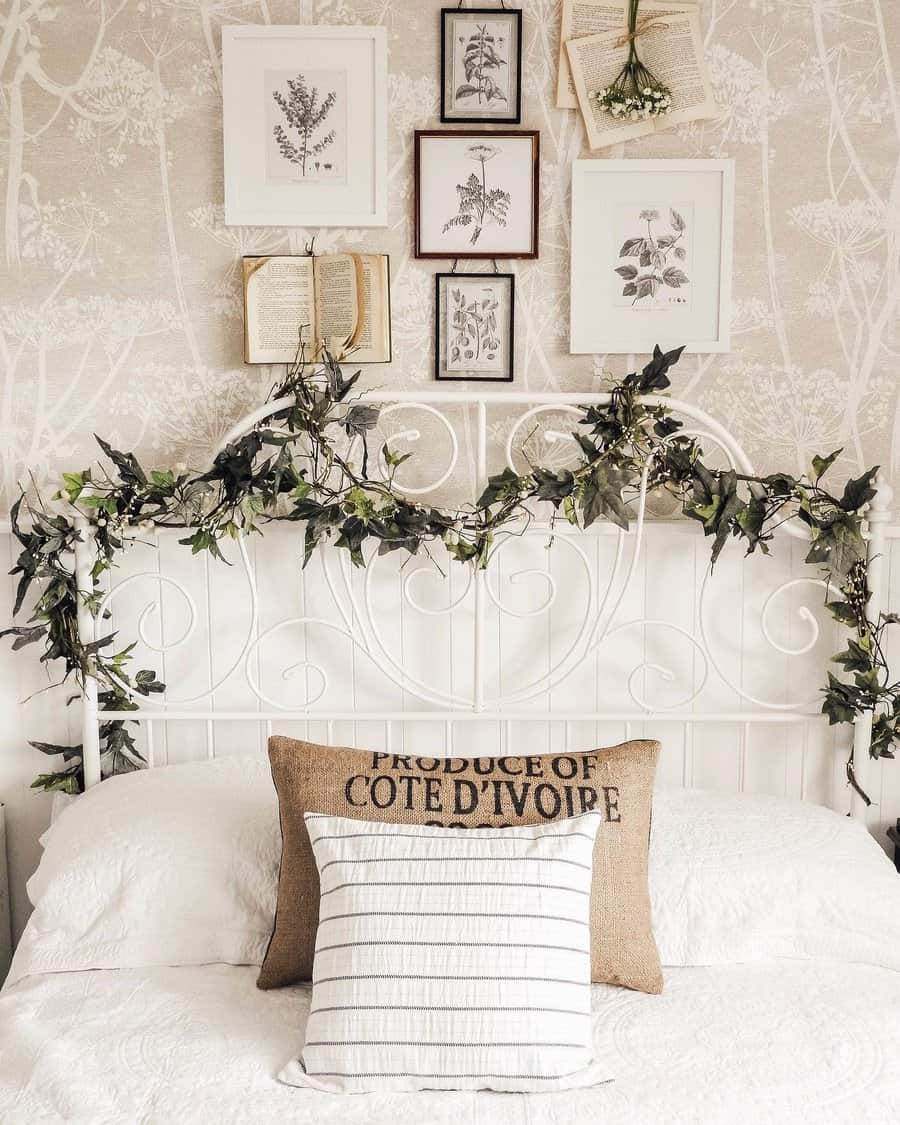 White bed with decorative iron headboard adorned with greenery, framed botanical prints and open book hang above, neutral-toned pillows