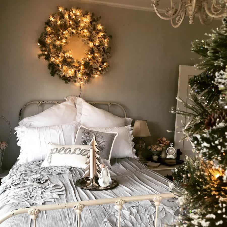 Cozy bedroom with a metal bed, decorative pillows, and a lit wreath on the wall. A small Christmas tree is in the foreground.