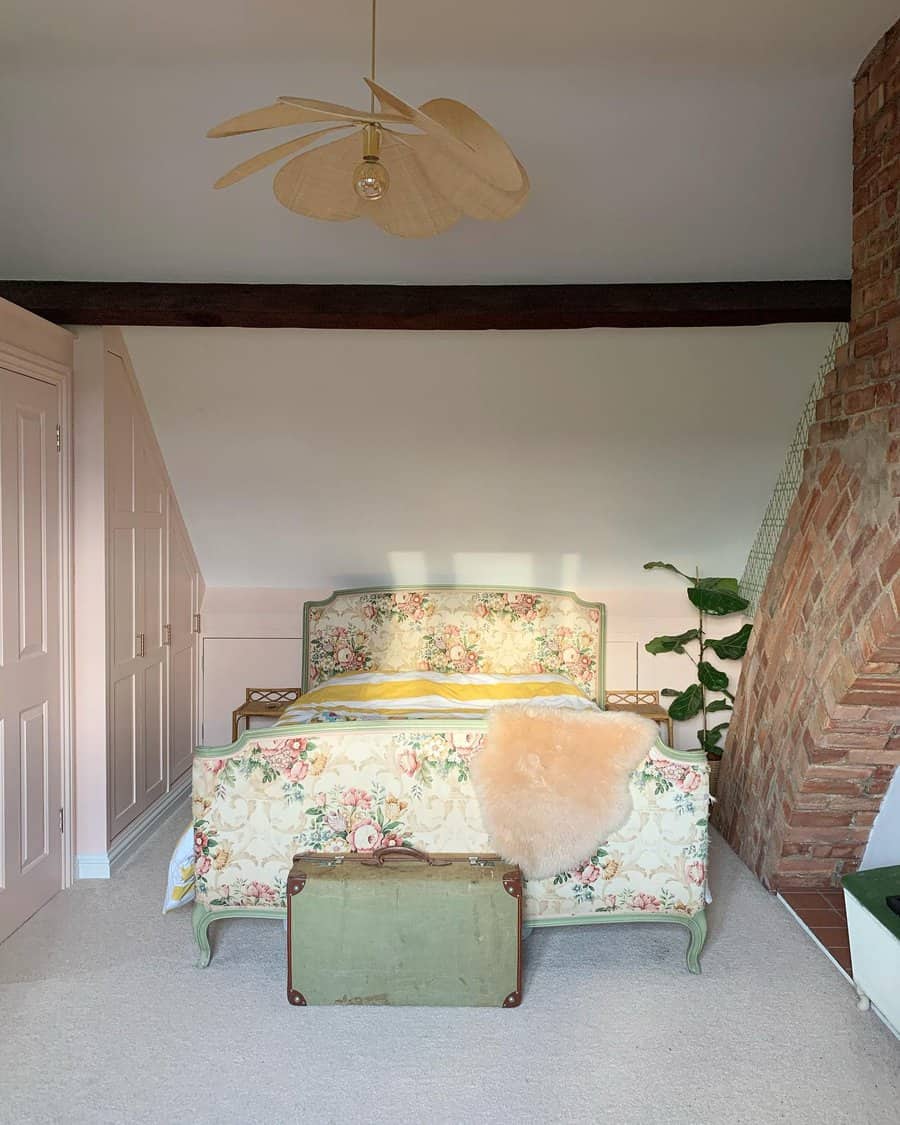 A cozy bedroom with a floral-patterned bed, wooden beams, a unique ceiling light, a plant, and a vintage suitcase at the foot of the bed