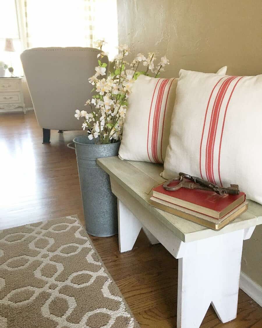 Charming cottage-style entryway with a rustic wooden bench, striped pillows, vintage books, and a metal bucket filled with delicate white flowers