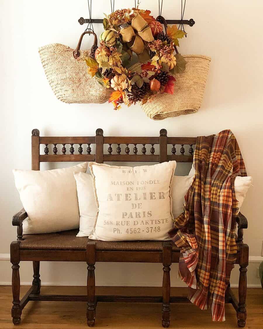 Wooden bench with white pillows and a plaid blanket; above, a basket holds autumn leaves and a wreath; cozy and rustic decor