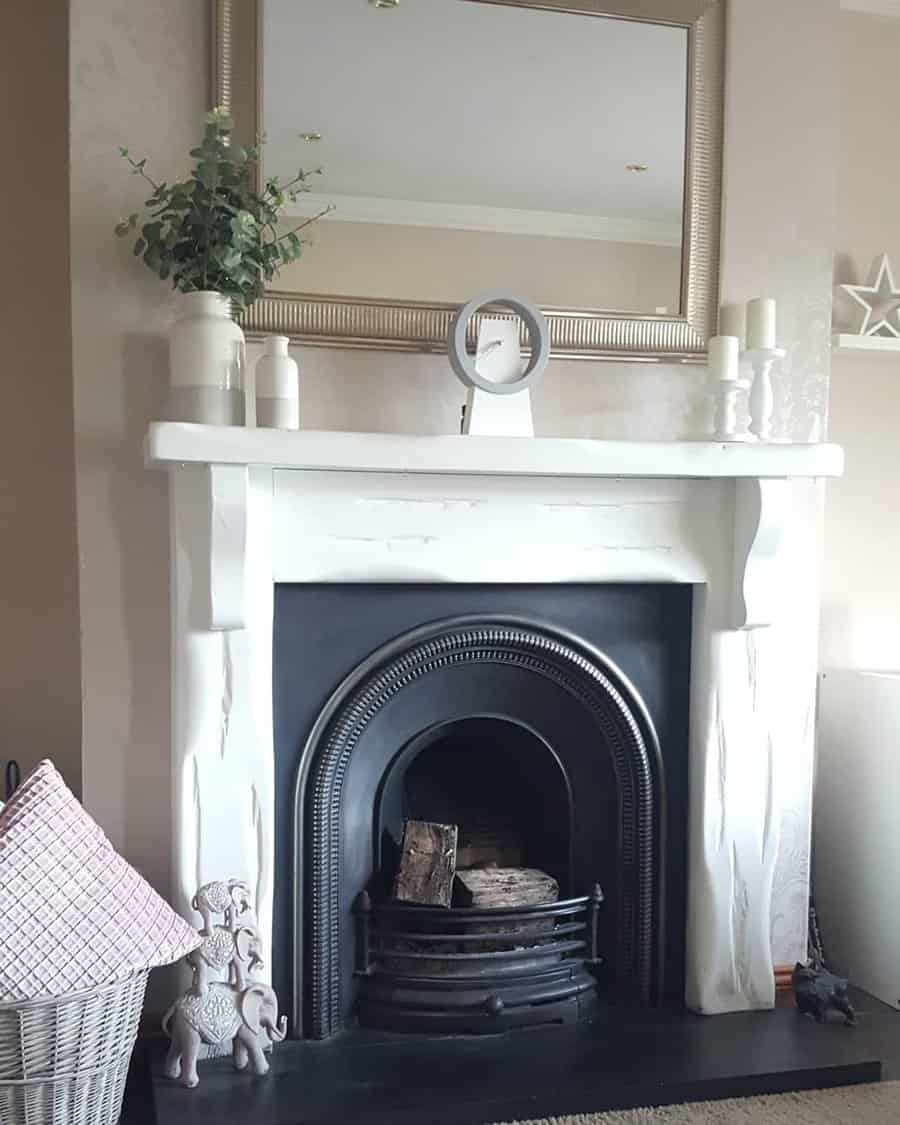 White mantelpiece with a mirror, candles, and decorative items above a black fireplace with logs; nearby are a basket and statues