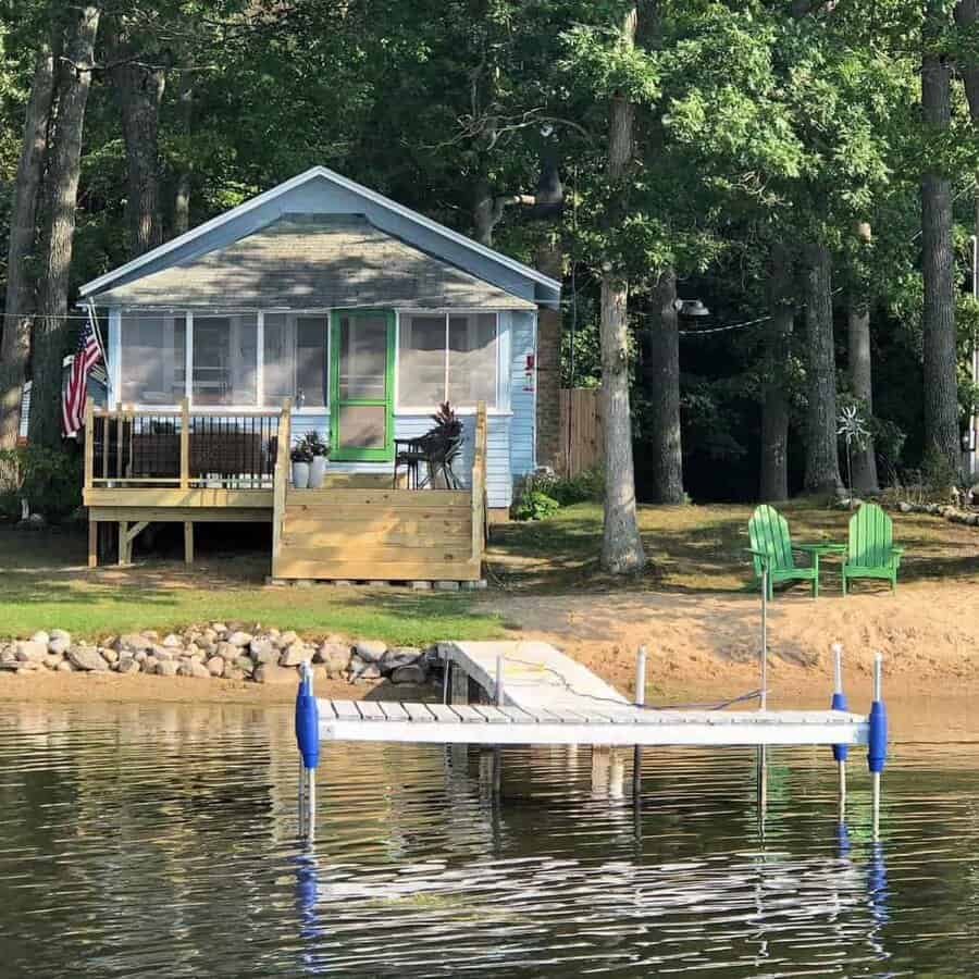 Small lakeside cabin with a dock, surrounded by trees and two green chairs on shore