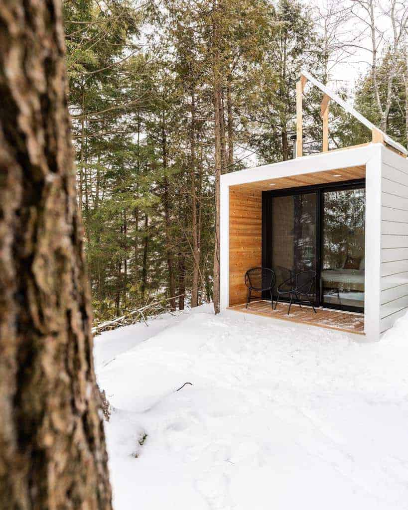 Modern cabin with glass doors, patio, and chairs in snowy forest setting