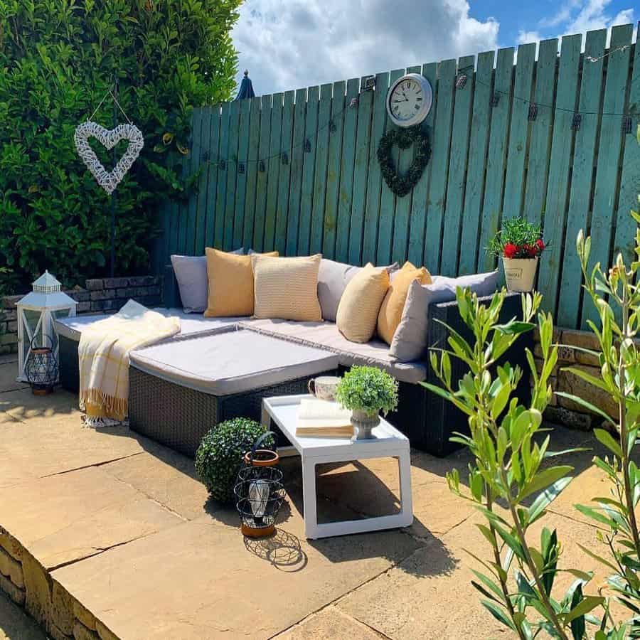 Cozy outdoor patio with a gray sectional, yellow pillows, a small white table, plants, and a turquoise fence adorned with decor