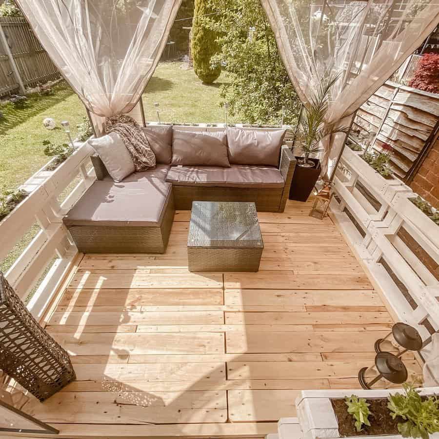 Cozy patio with a pallet sofa, cushions, and glass-topped table on a wooden deck, complemented by sheer curtains and plants framing the view of the garden