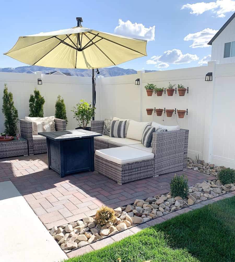 Patio with wicker furniture, a large umbrella, potted plants on a white fence, and a fire pit, surrounded by pebble landscaping