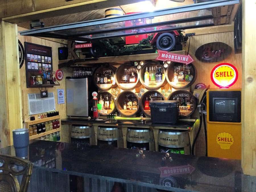 Bar with circular bottle shelves, neon signs, barrels labeled "Federalist," and vintage decor on wooden walls