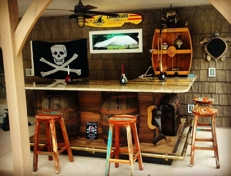 Outdoor tiki bar with wooden stools, pirate-themed decor, skull and crossbones flag, surfboard sign, and barrels under granite counter