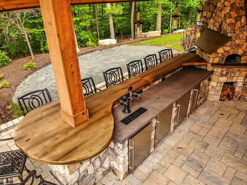 Outdoor bar with wood and stone design, metal stools, and a fieldstone counter surrounded by trees and a stone patio