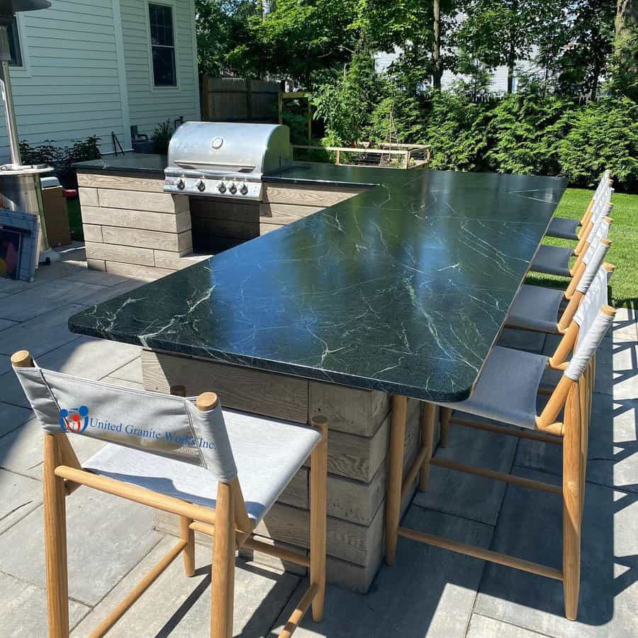 Outdoor kitchen with a black granite countertop, bar seating, and a stainless steel grill, set on a stone patio with greenery