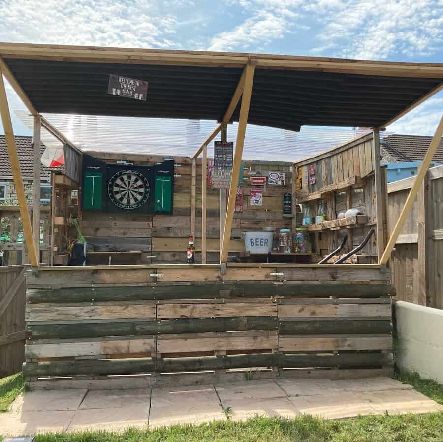 Wooden outdoor bar with a dartboard, signs, and a corrugated roof, surrounded by a fence, set on a grassy yard