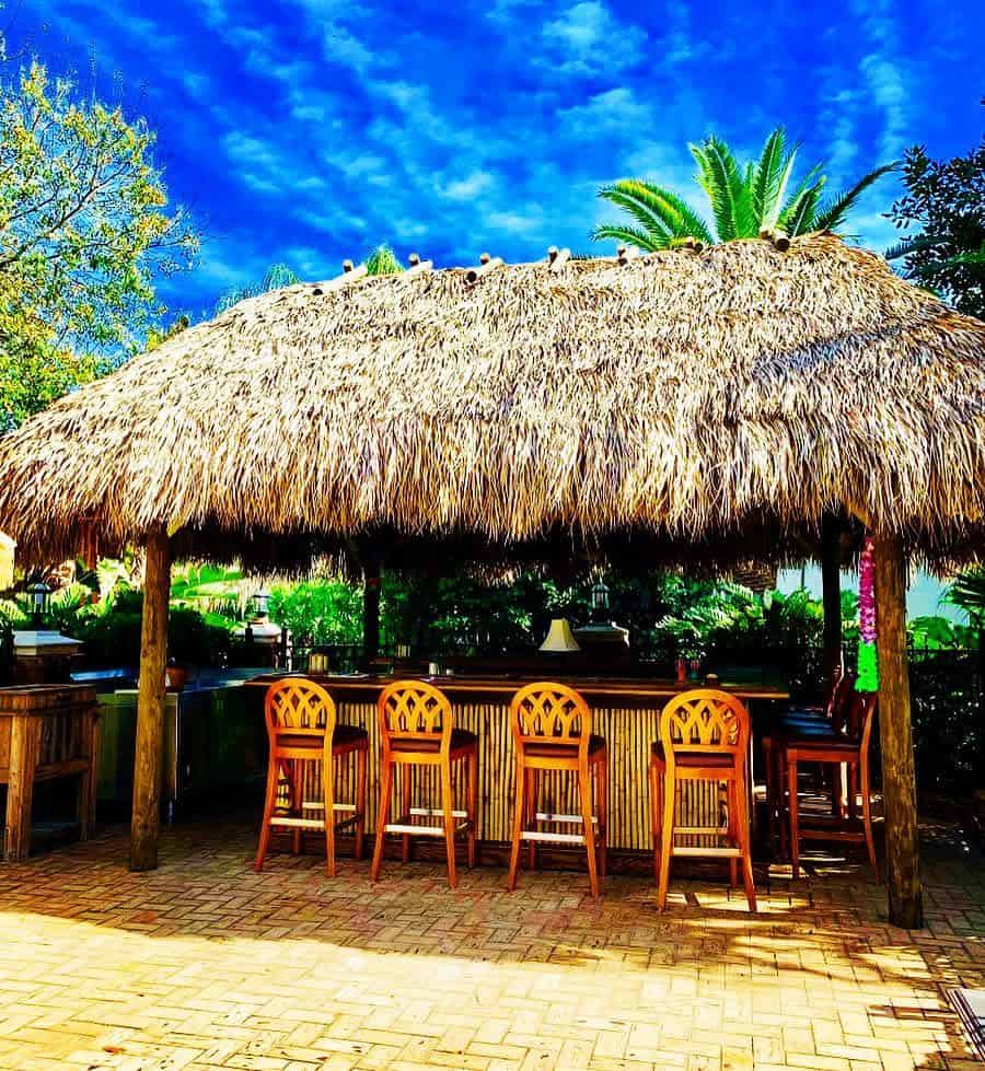 Outdoor tiki bar with a thatched roof and wooden stools, surrounded by greenery, under a bright blue sky