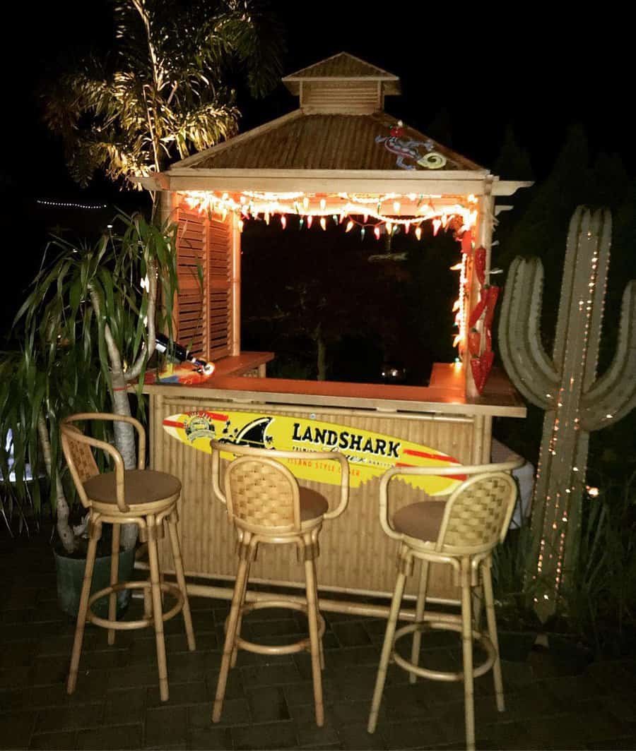 Outdoor tiki bar at night with stools, string lights, a large cactus, and a "Landshark" surfboard sign