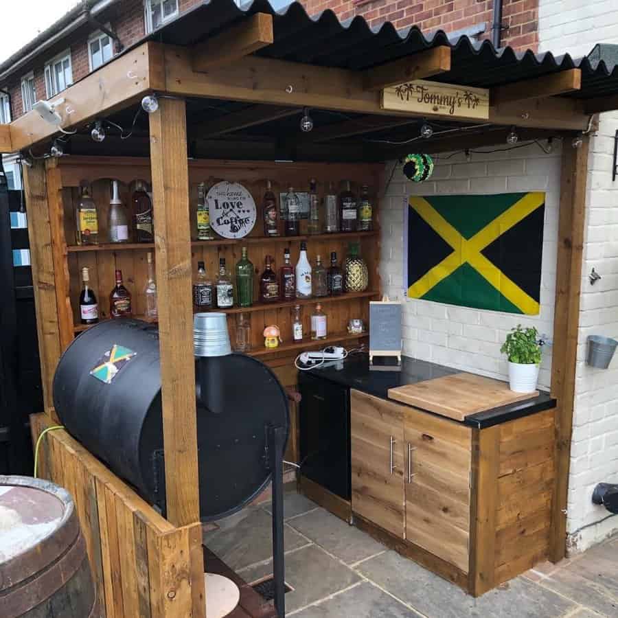 Outdoor bar with wooden shelves, various bottles, a barbecue grill, and a Jamaican flag hanging on the wall