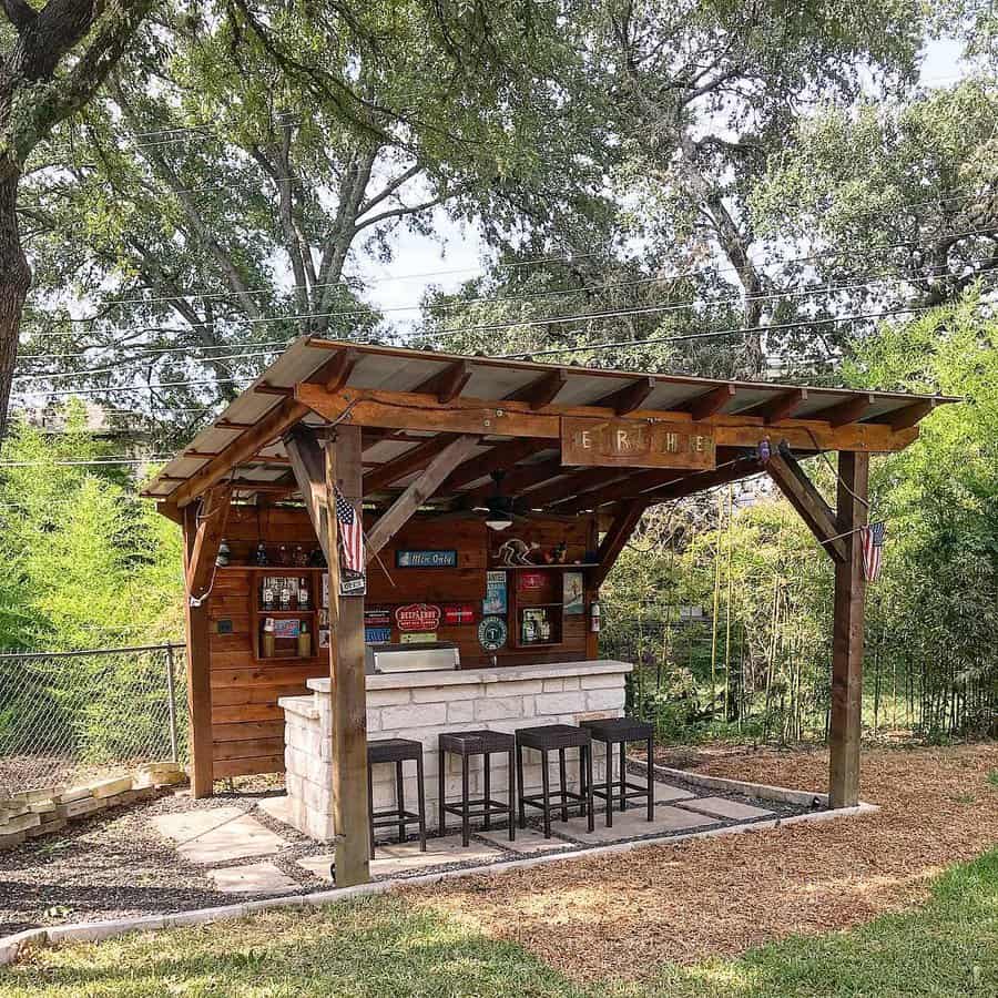 Rustic backyard bar with a wooden pergola, stone counter, and barstools, surrounded by greenery for a cozy outdoor retreat