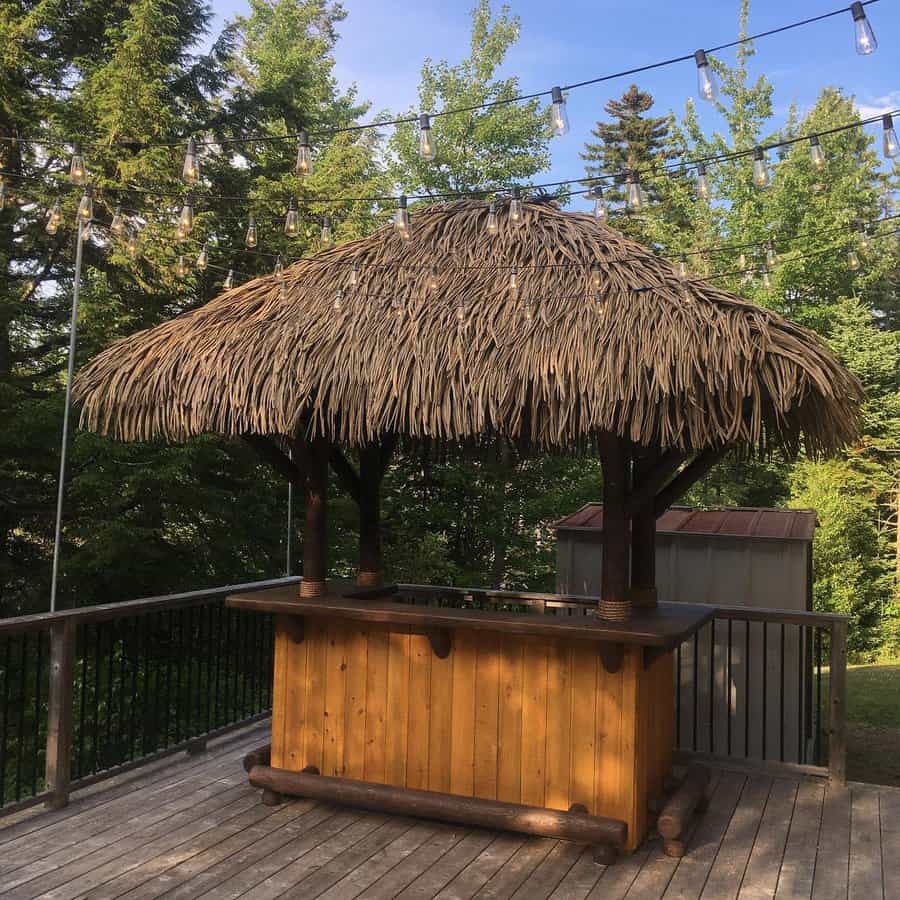 Tiki bar with a thatched roof on a wooden deck, surrounded by string lights and lush green trees