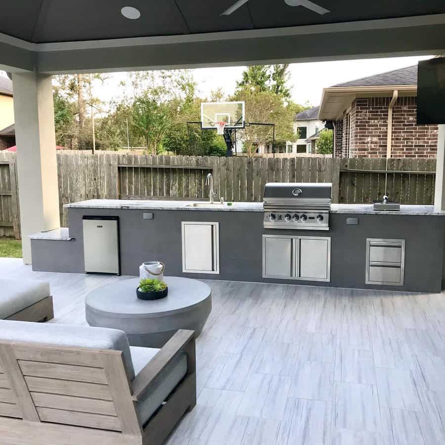 Outdoor kitchen with a grill, sink, and mini fridge; gray patio seating and table in front with a basketball hoop in the background