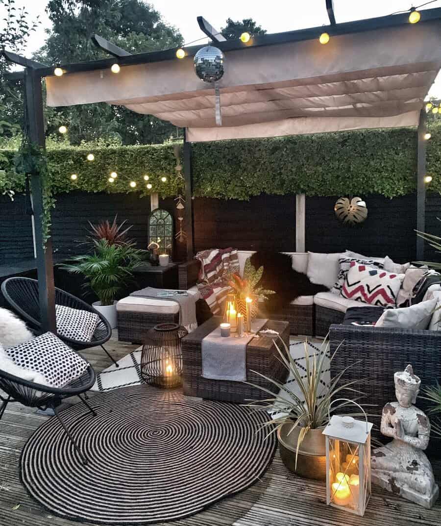 Cozy outdoor patio with wicker furniture, cushions, plants, and soft lighting. A round rug and lanterns enhance the inviting atmosphere.