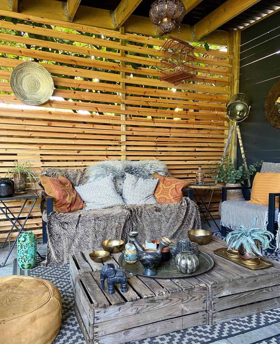 Cozy outdoor patio with a sofa, pillows, fur throw, wooden crate table, ornate decor, and slatted wooden wall backdrop