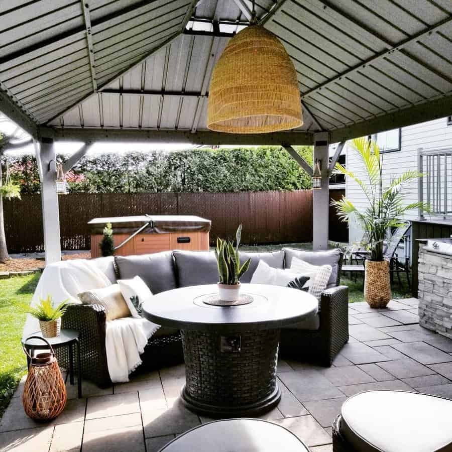 A cozy outdoor patio with a round wicker sofa, pillows, a central fire pit table, wicker lanterns, and potted plants under a gazebo