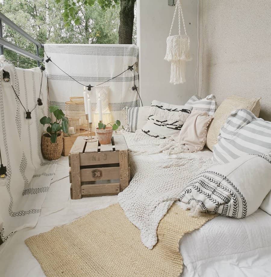 Boho-style small balcony with cozy floor seating, layered cushions, neutral-toned textiles, a wooden crate table, plants, and string lights