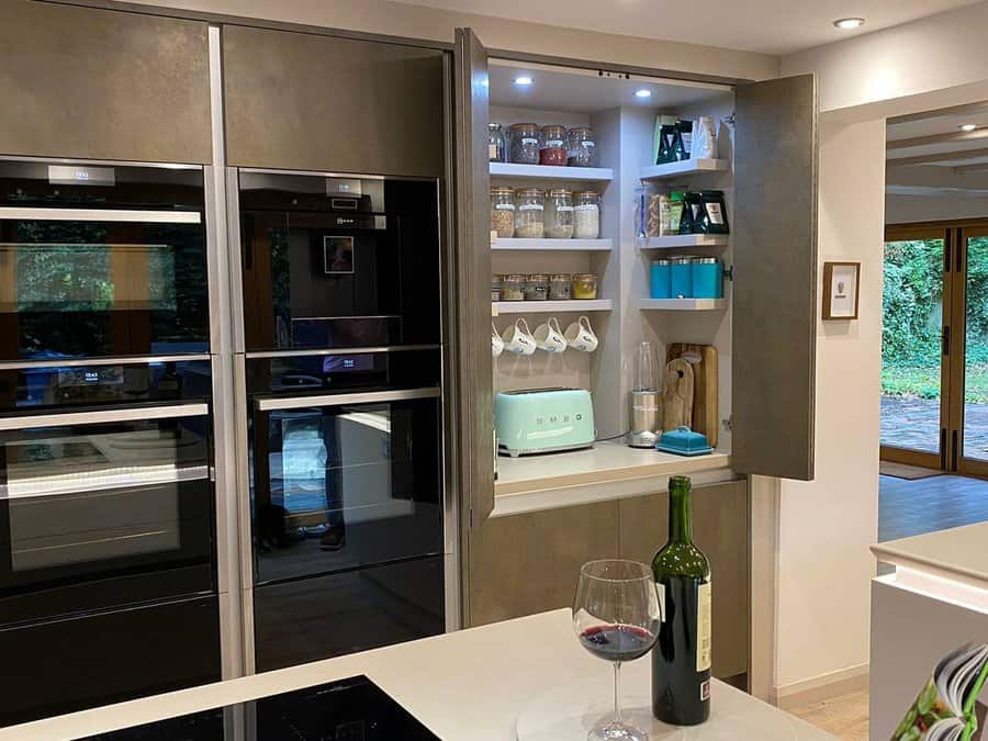 Modern kitchen with a hidden pantry cabinet featuring shelves with jars, mugs, and appliances, blending sleek design with smart storage