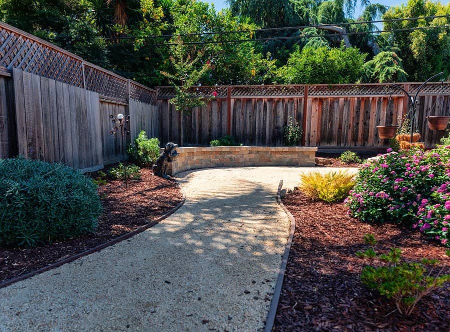 Backyard landscaping with a gravel pathway, vibrant flower beds, wooden fence, and a curved stone retaining wall for a natural look