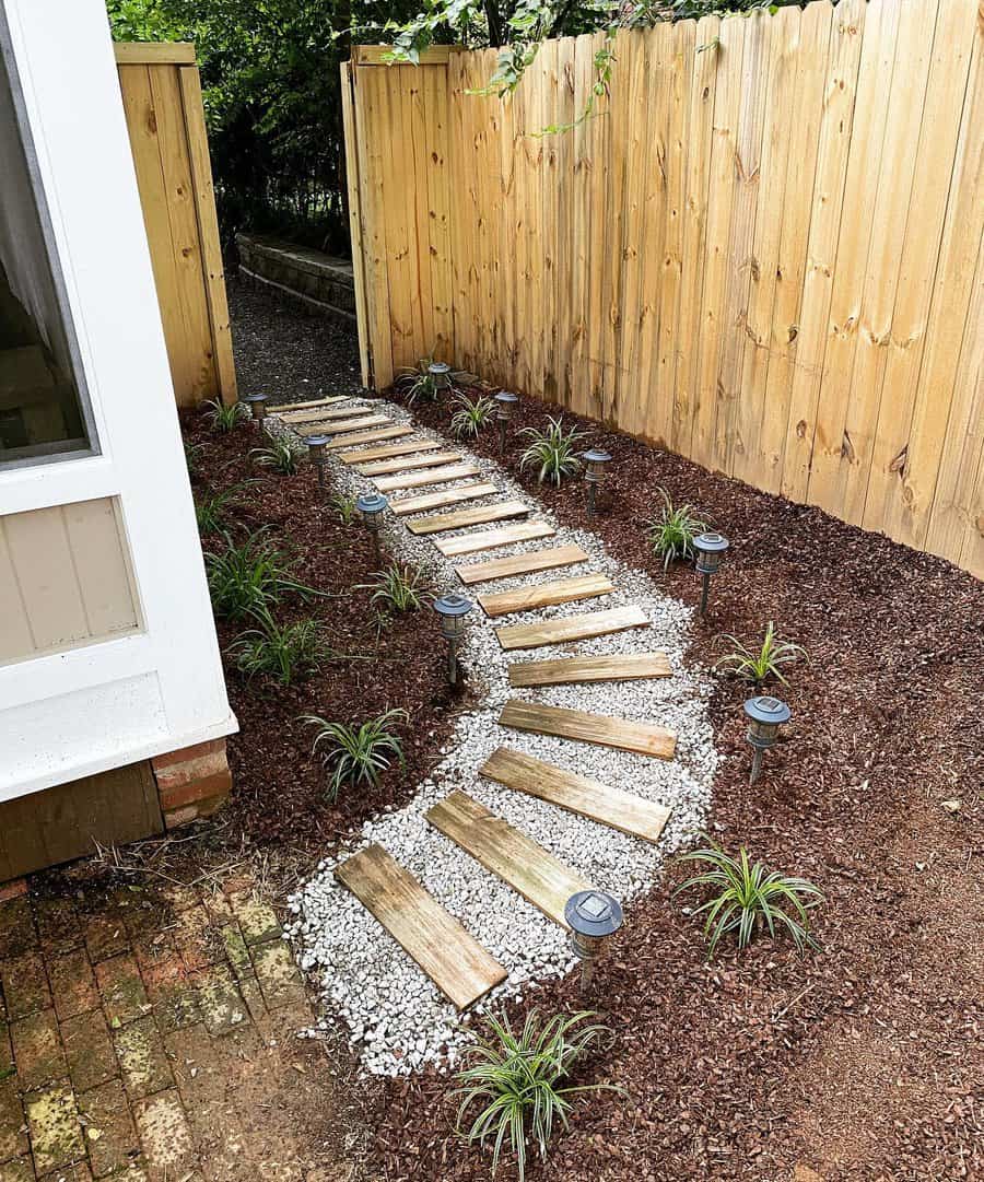 Side yard with a wooden and gravel pathway, bordered by mulch and small plants, complemented by solar pathway lights and a wooden fence