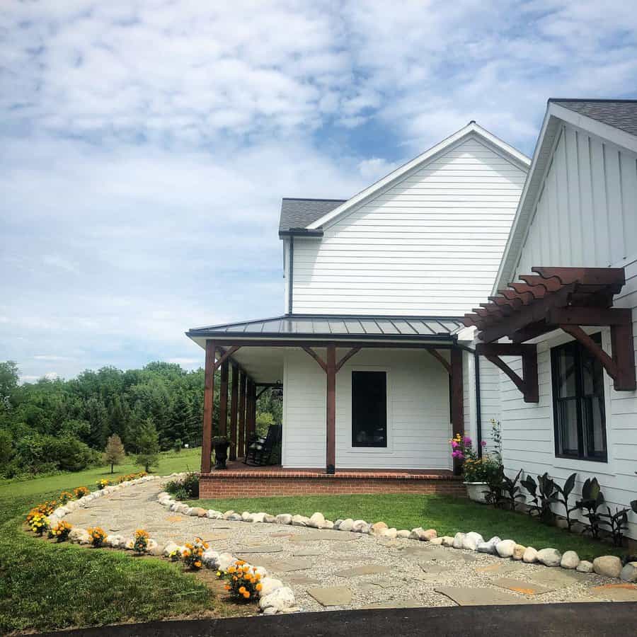 Farmhouse landscaping with a stone pathway lined with bright marigolds and rocks, leading to a porch with wooden beams and a pergola