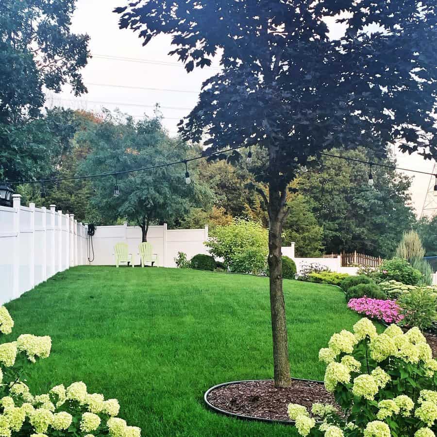 Spacious backyard with lush green grass, blooming hydrangeas, a tree with string lights, and two Adirondack chairs by a white fence