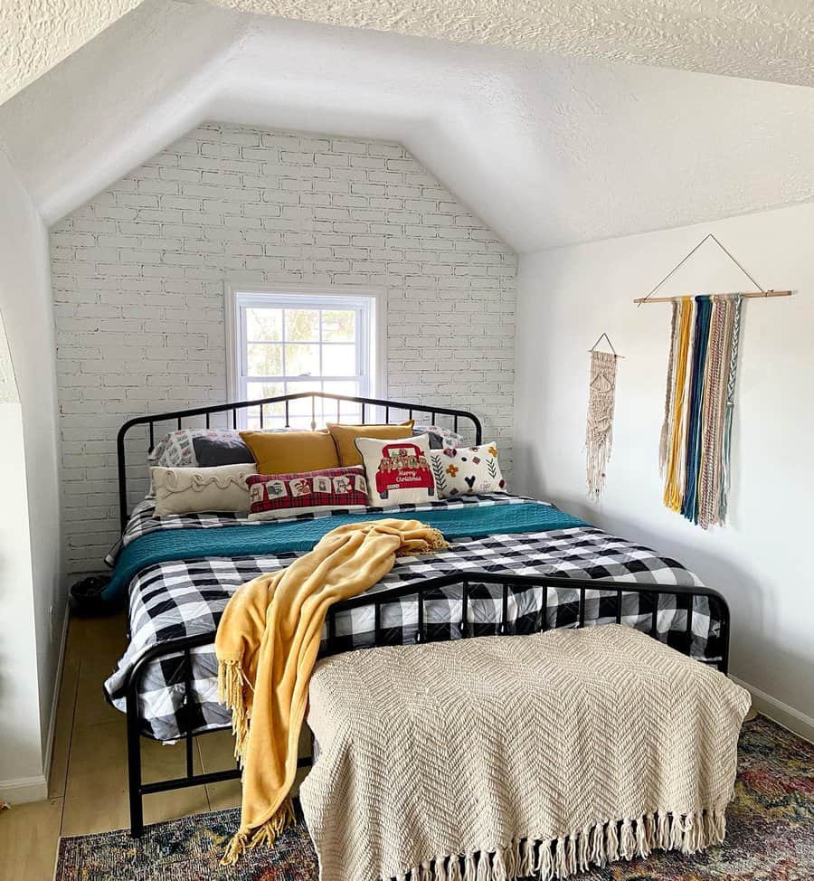 Cozy attic bedroom with a black metal bed frame, buffalo plaid bedding, and colorful boho accents. A white brick wall adds rustic charm.