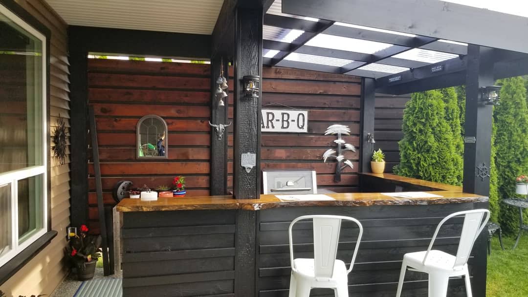 Outdoor bar setup with a rustic wooden counter, bar stools, and BBQ sign surrounded by greenery and decorative plants