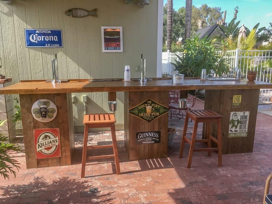 Outdoor wooden bar with stools, decorated with vintage beer signs and drink taps, set on a brick patio with plants and trees nearby