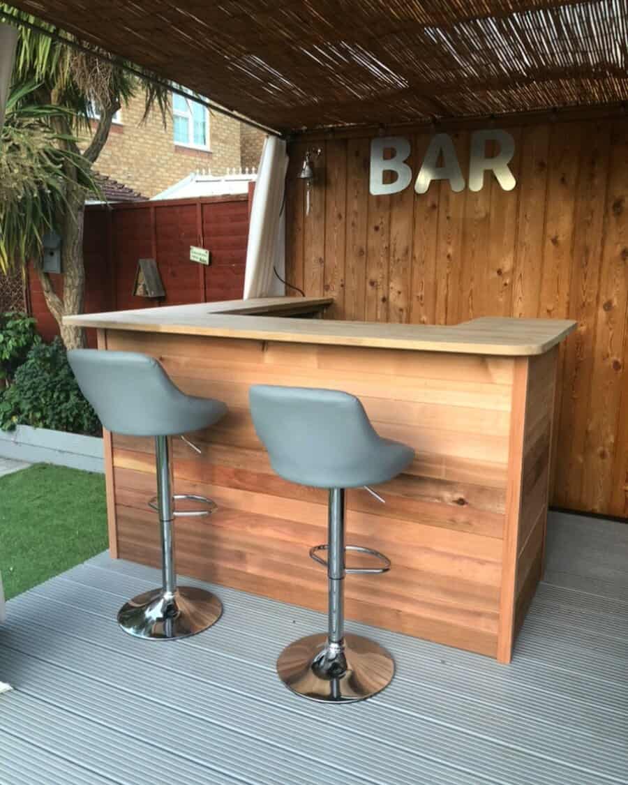 Outdoor wooden bar with two gray bar stools under a thatched roof, featuring a "BAR" sign on the back wall