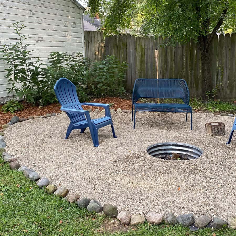 Backyard fire pit area with gravel ground, surrounded by stones, blue chair and bench sit nearby, fence and trees in the background