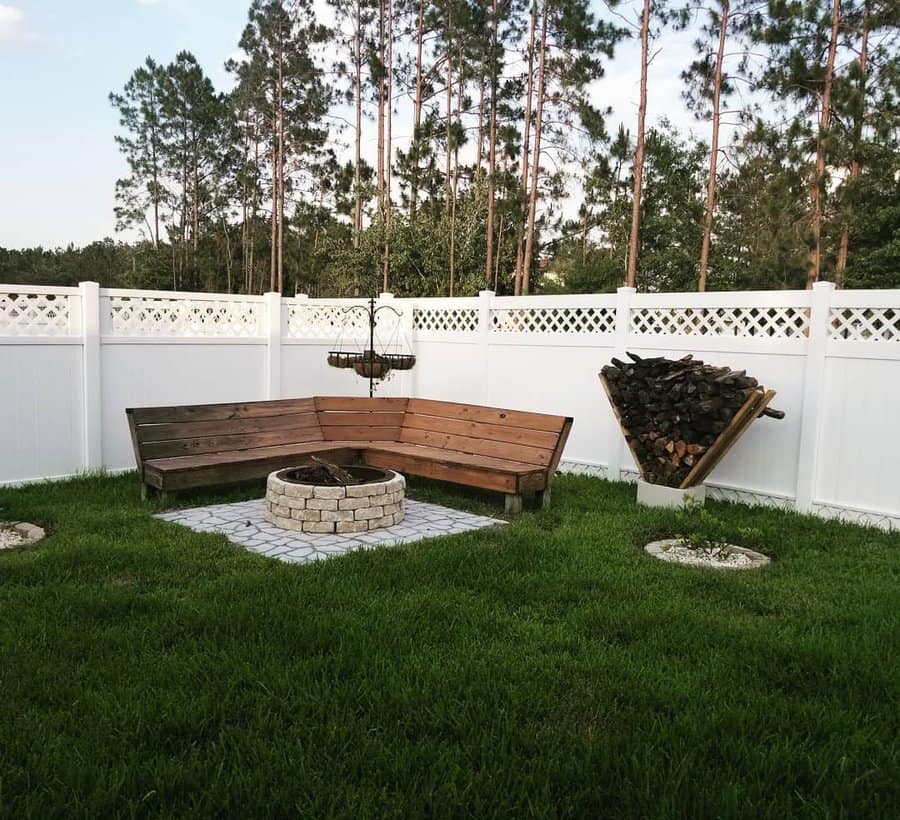 Lush green backyard with wooden benches around a stone fire pit, a white fence, stacked firewood, and tall trees in the background