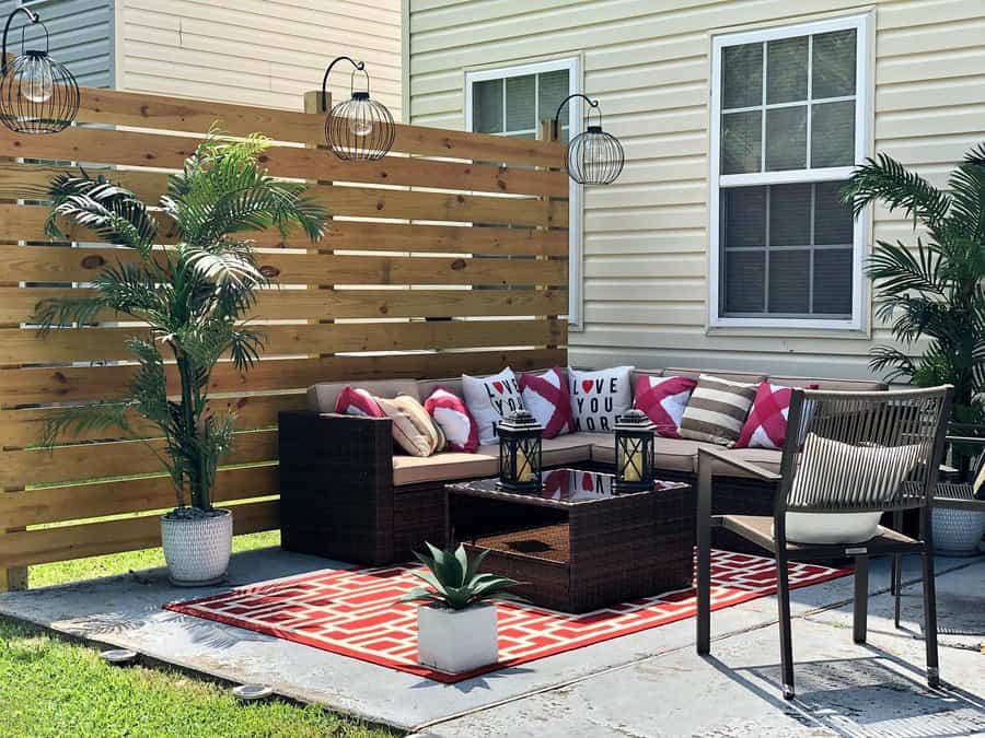 Cozy patio with a sectional sofa, red and white pillows, potted plants, and two lanterns on a coffee table, surrounded by wooden fencing